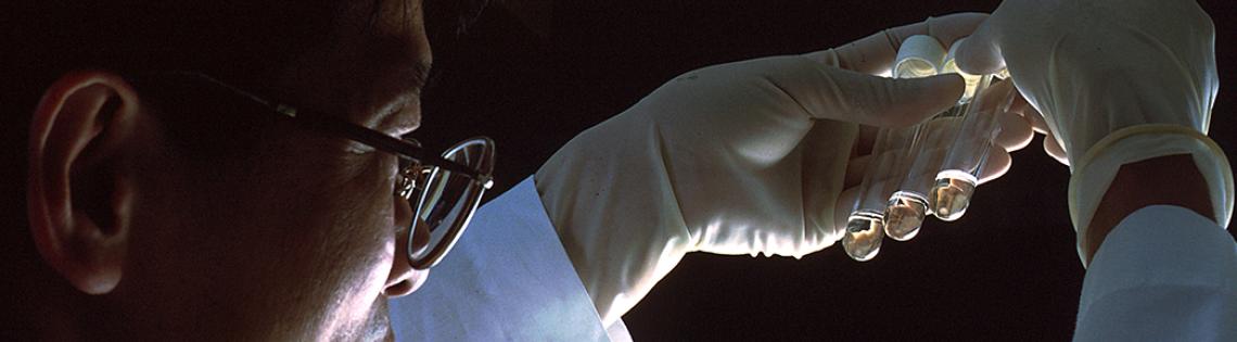 An NCI scientist in white lab coat and gloves holds up 3 test tubes.