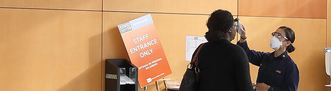 Panoramic view of hospital lobby with uniformed nurse taking temperature of employee entering building.