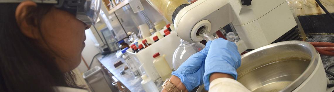 A female trainee wearing safety goggles and blue gloves does an experiment at a lab workstation, handling liquid over a large bowl. 