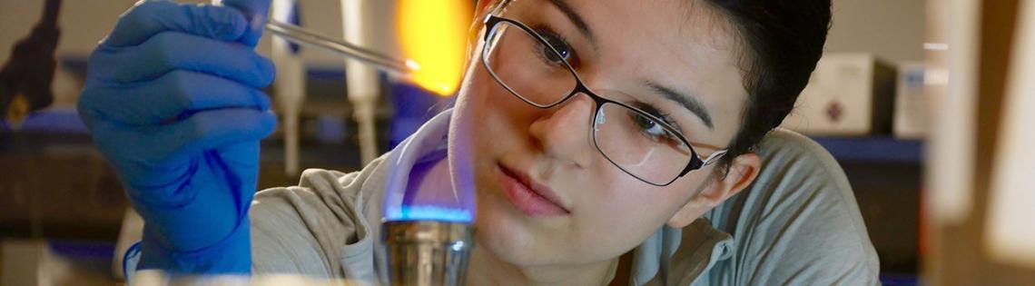 Closeup image of woman with lab-gloved hand holding an eyedropper over an open flame.