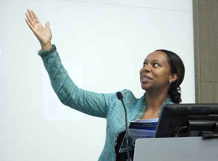 Lash gestures up toward slide as she speaks from behind podium.
