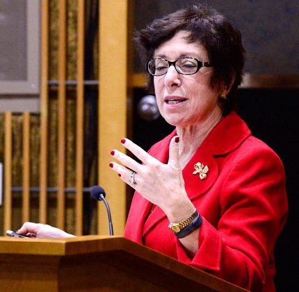 Birnbaum, dressed in red, speaks at podium.