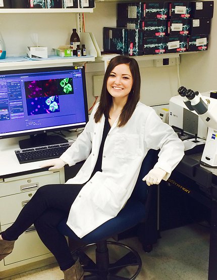 Stahl, in white lab coat, sits in front of computer monitor, next to microscope.