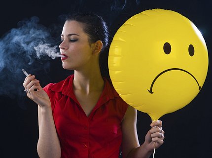 A woman blows smoke cloud and holds cigarette in one hand, a yellow, frowning-face balloon in the other