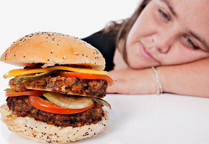 A girl rests her head on the table looking longingly at a large burger