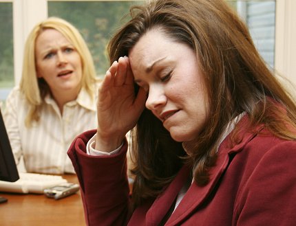 A woman sits holding her head as another woman looks on with concern.