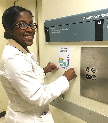 Holshue, in a white lab coat, stands in front of Take the Stairs sign next to elevator in the Clinical Center