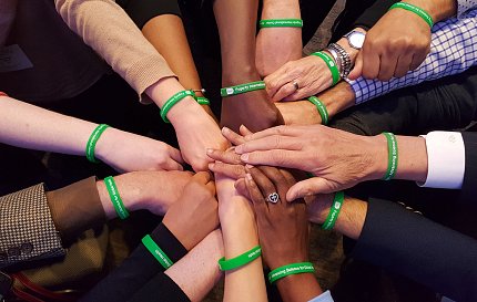 A dozen arms extended, each wearing a green wristband, with palms down, hands stacked