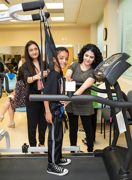 A smiling child wears a safety harness while walking on a treadmill in the Clinical Center's Rehab Clinic. 