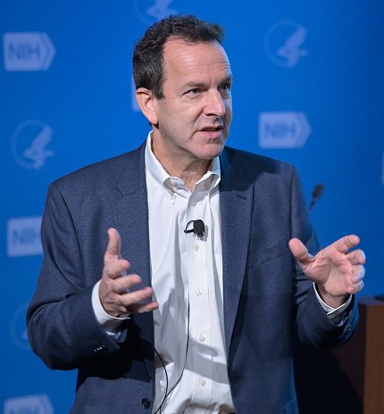 Salzberg stands on stage in Lipsett, hands open, in front of blue NIH backdrop.