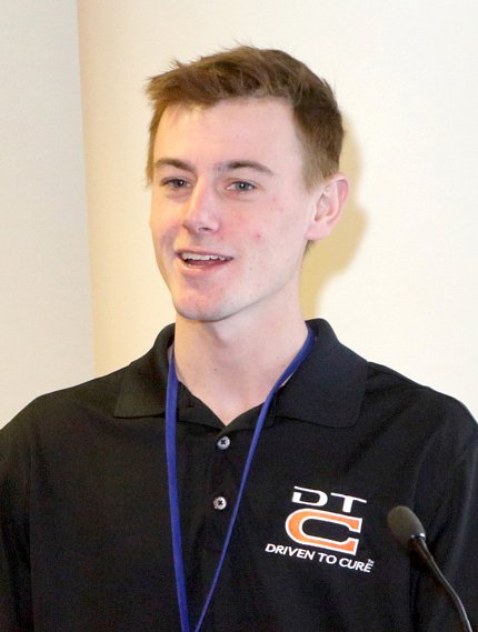 Head and shoulders of Lee, in Driven to Cure logo shirt, smiling at podium