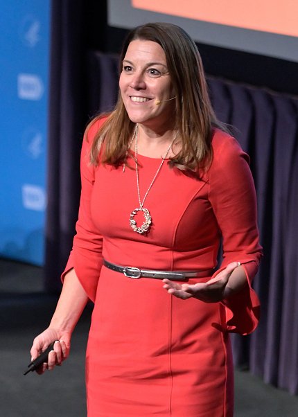 Karin Hurt in red dress stands on stage with hand extended.