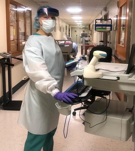 Lola Saidkhodjaeva in scrubs and a face shield stands by a computer station in a Clinical Center hallway.