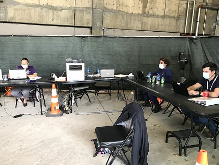 Several workers in protective masks sit at a series of tables put together to form an "L" shape. 