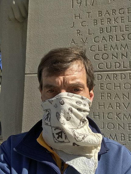 Professor Ewing, wearing a cloth mask, stands in front of pylon etched with names of World War I victims