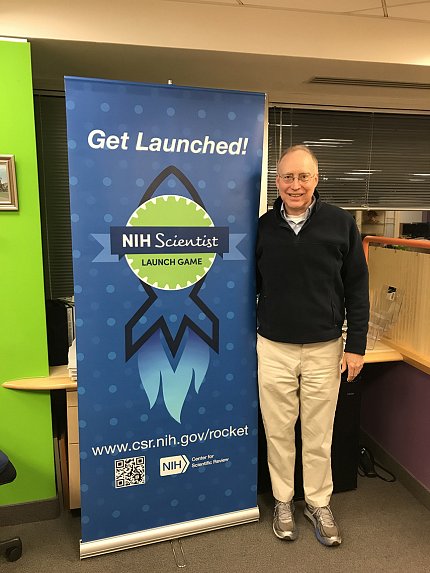 Lucketts stands to the left of a banner that reads, "Get Launched! NIH Scientist Launch Game" along with the web address for the game. 
