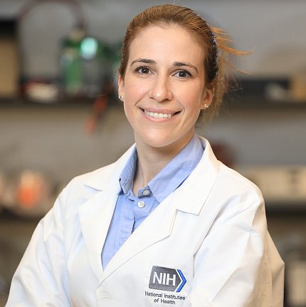 A smiling Sadtler, in white lab coat with NIH logo, standing in the lab