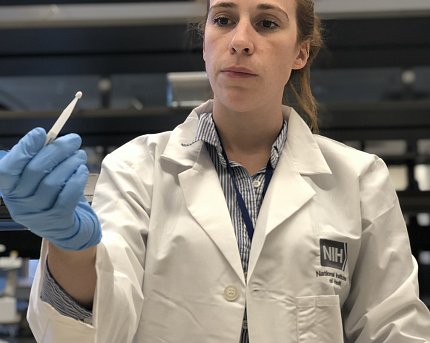Sadtler, in white lab coat and blue gloves, holds up a lancet used to prick the finger to collect a blood sample.