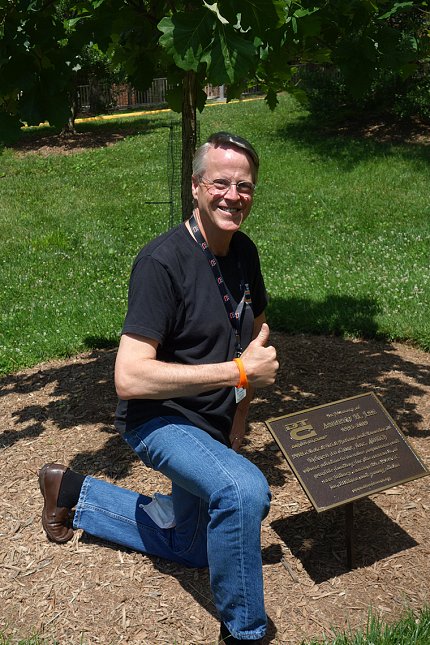 Bruce Lee gives thumbs up as he kneels, smiling next to the tree and memorial plaque honoring his late son, on the south lawn of the Clinical Center.