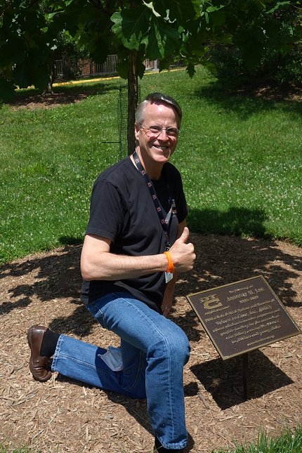 Bruce Lee gives thumbs up as he kneels, smiling next to the tree and memorial plaque honoring his late son, on the south lawn of the Clinical Center.