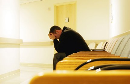 A young man sits, head in hands, at end of row of yellow chairs in hospital.
