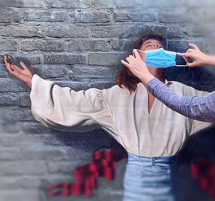 Person posed with outstretched arms, back to a wall, while a pair of hands places a mask over the person's nose &amp; mouth
