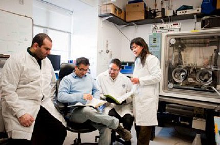 Three members in white coats stand around Bharti while Bharti reads from a notebook in his lab