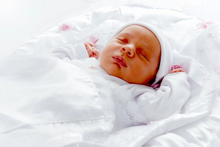 Infant dressed in white with white blanket lies on her back sleeping