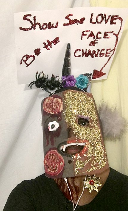 Person wearing hand-made face mask with sign overhead reading "Show Some Love. Be the Face of Change"