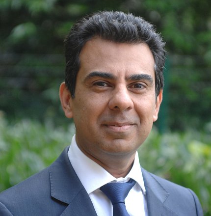 Headshot of Shamez Ladhani in a suit standing outside in front of trees and shrubs