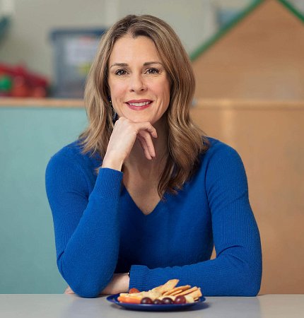 Dr. Bailey sits, resting chin on hand, with a plate of fruit, crackers on table in front of her.
