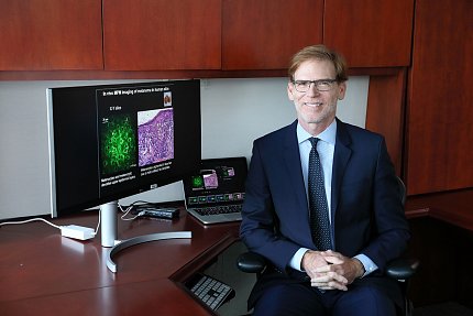 Tromberg in suit and tie sits at a deck with a computer monitor and a laptop beside him.