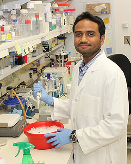 Dr. George stands in white lab coat by shelves of equipment and supplies with his hands around a red bowl filled with ice