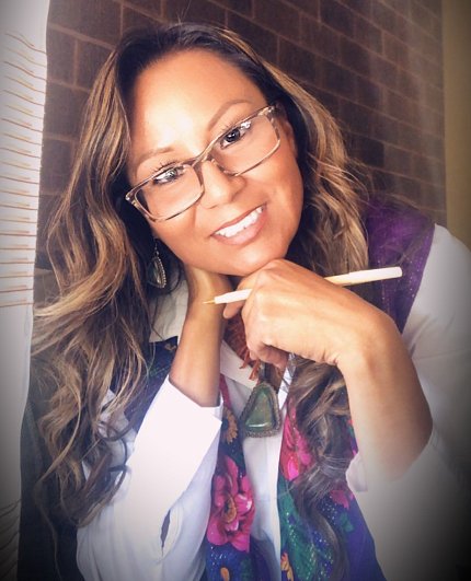 Casual photo of Segay in colorful scarf; she has elbows on the table, leaning chin on hand, holding a pen.