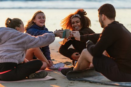 Four adult friends sit in a circle on the beach clinking coffee mugs