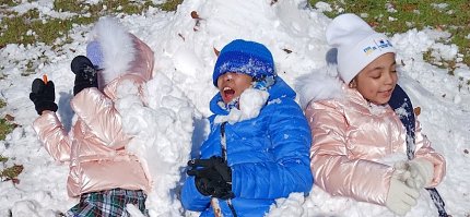 Wearing a blue puffer jacket, hat and gloves, Caesar laughs as he rolls around in the snow with his 2 sisters.