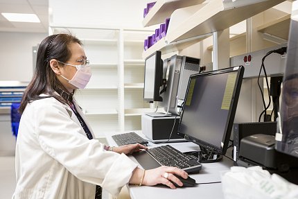 Woman in lab coat stands at computer screen while using mouse attachment.