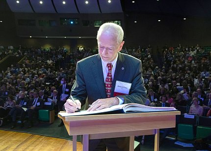On stage with audience behind him, Youle writes in a ledger on a podium.