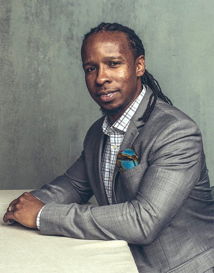Dr. Kendi sitting in a grey suit, hands folded on table, in front of grey wall