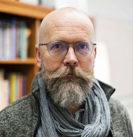 Headshot of Thomas, grey coat and scarf, in front of bookcase