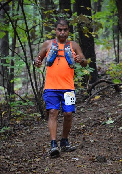Bhattacharyya in orange shirt, blue shorts and backpack running on a trail through the woods