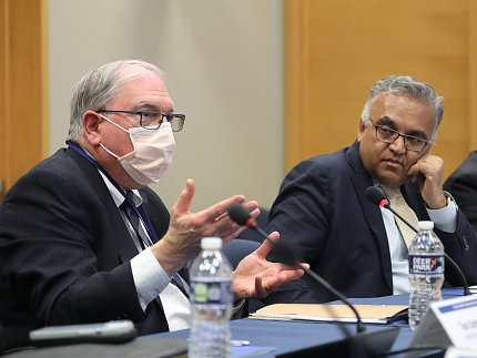 Seated at a conference table, Tabak (l) gestures toward Jha (r).