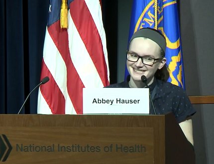 Hauser stands smiling, talking into mic at podium