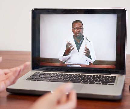 A doctor talks through laptop screen, pair of hands near keyboard of person listening 