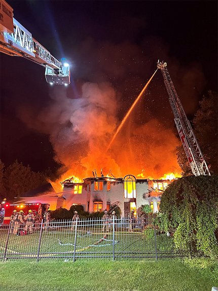 Flames and smoke engulf a large house in Maryland as a NIHFD ladder hovers over, with a fireman up top spraying water from a hose