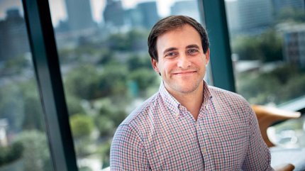 McLeod wears a red, blue and white plaid shirt and smiles at the camera. Behind him is a window with a cityscape beyond.