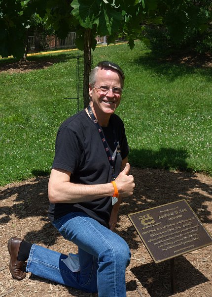 A smiling Bruce Lee giving a thumbs up kneels by plaque on lawn on the Clinical Center's south side.
