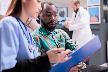 Asian woman wearing scrubs and stethoscope holding hospital chart towards a Black man