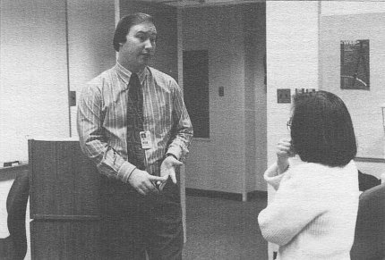 Black-and-white photo of McLean, wearing a striped shirt and dark tie, speaking to a woman who is partially out of frame