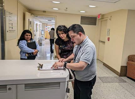 Van Berg, in a gray polo shirt, points to a clipboard on a desk as Wolek and another woman look on.
